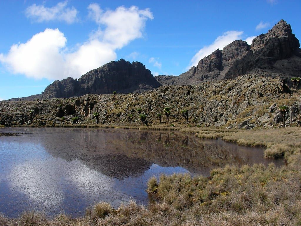 Rugged rocky peaks and blue skies reflected in a calm alpine tarn on Mount Kenya.