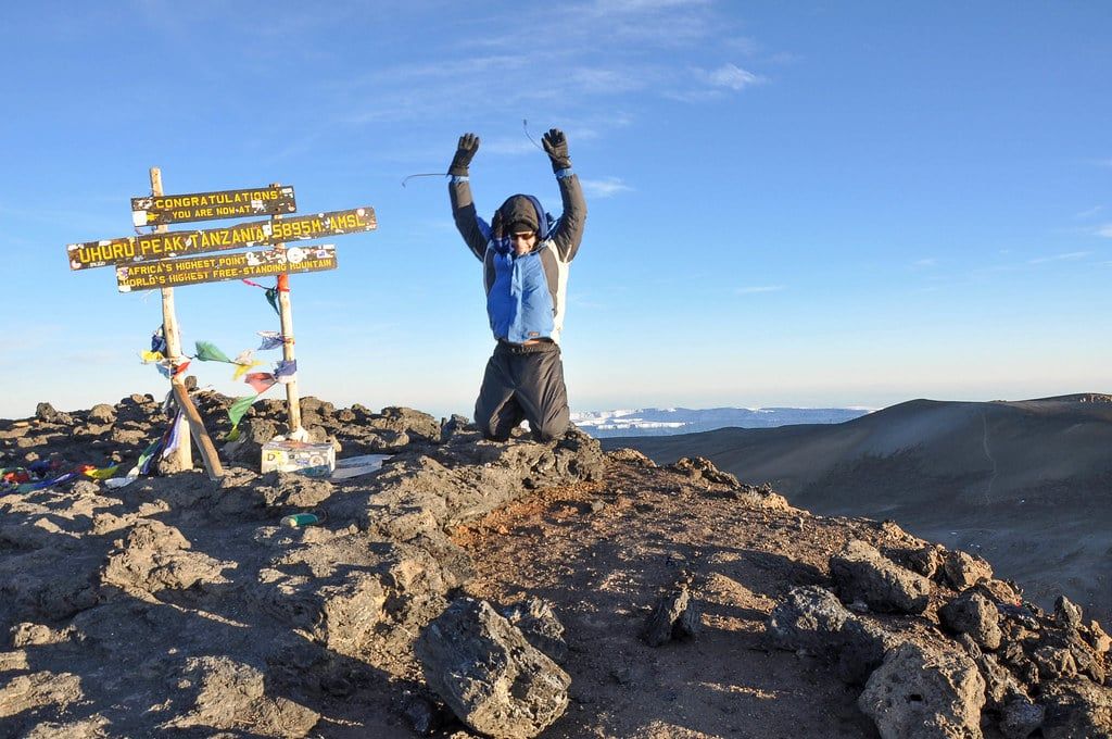 A climber celebrates next to the wooden summit sign marking Uhuru Peak on Mount Kilimanjaro.