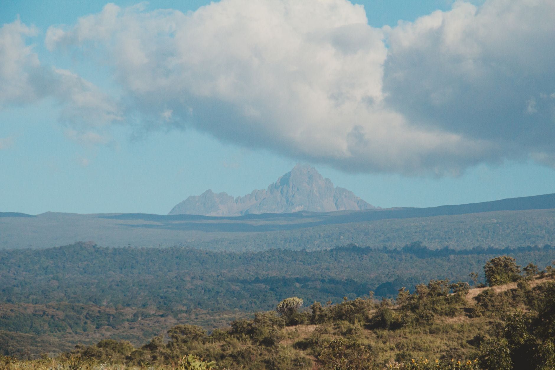 The jagged peaks of Mount Kenya rise in the distance above rolling forested hills beneath a cloud-filled blue sky.